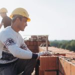 Construction worker laying bricks outdoors in sunny weather with lush greenery in background.