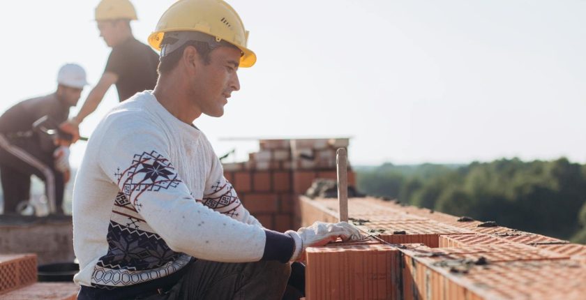 Construction worker laying bricks outdoors in sunny weather with lush greenery in background.