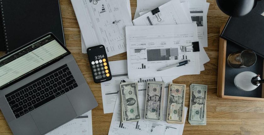 Overhead view of financial documents, cash, and technology on a wooden desk.
