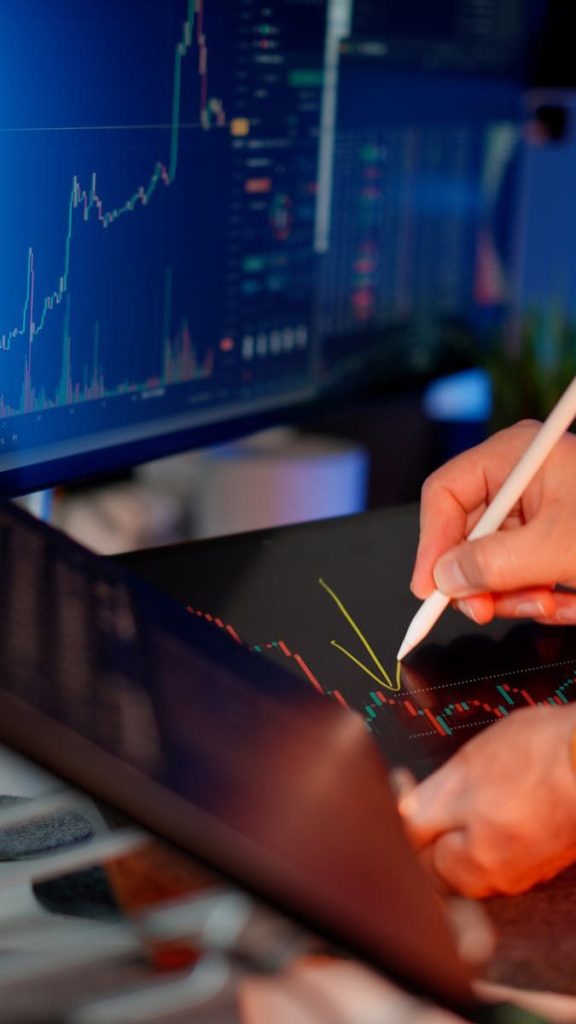 Person using a stylus on a tablet for stock market analysis, with charts on a monitor.