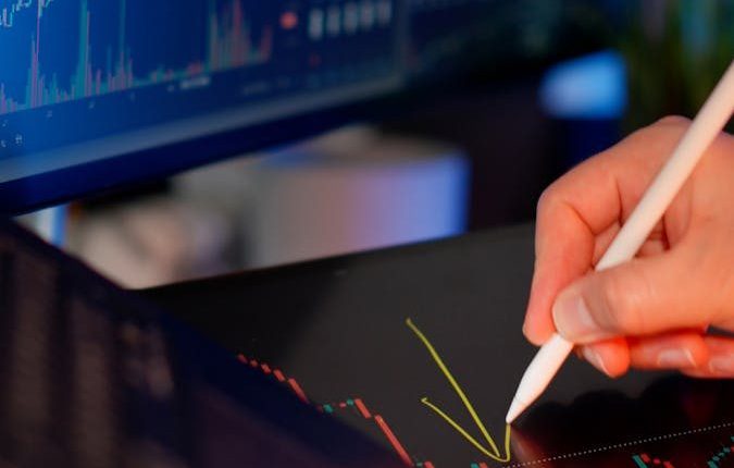 Person using a stylus on a tablet for stock market analysis, with charts on a monitor.