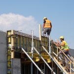 Two construction workers building a structure, embodying teamwork and safety under a clear blue sky.