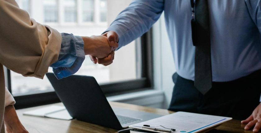 Crop unrecognizable coworkers in formal wear standing at table with laptop and documents while greeting each other before meeting